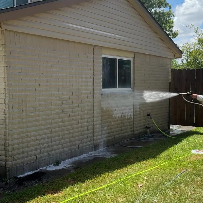 Water spraying along a tan brick wall below a window, with white suds collected along the base of the wall